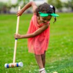 A child swings a croquet mallet toward a yellow ball on the lawn