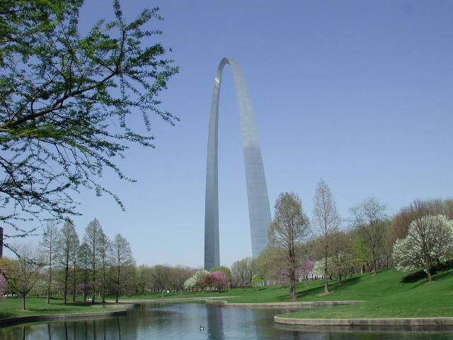 The Gateway Arch viewed through spring foliage from park grounds beside a water feature