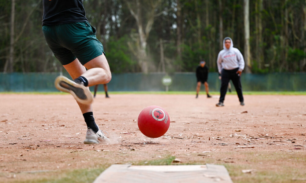 Real kickball player striking a red kickball on a dirt field during a tournament