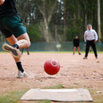 Real kickball player striking a red kickball on a dirt field during a tournament