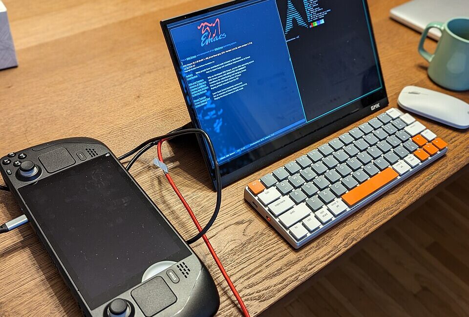 Real Steam Deck handheld on a desk beside an external screen and keyboard, illustrating the battery-powered gaming PC