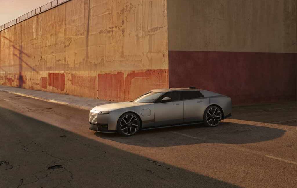Silver Jaguar Type 01 electric grand tourer parked beside an industrial wall at sunset.