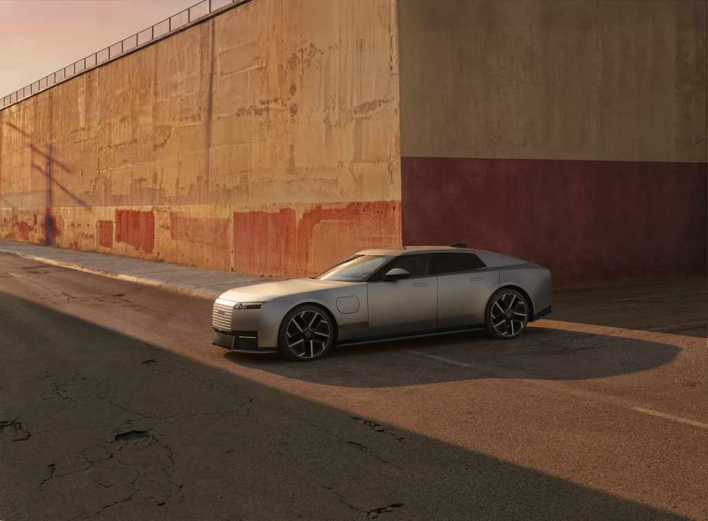 Silver Jaguar Type 01 electric grand tourer parked beside an industrial wall at sunset.