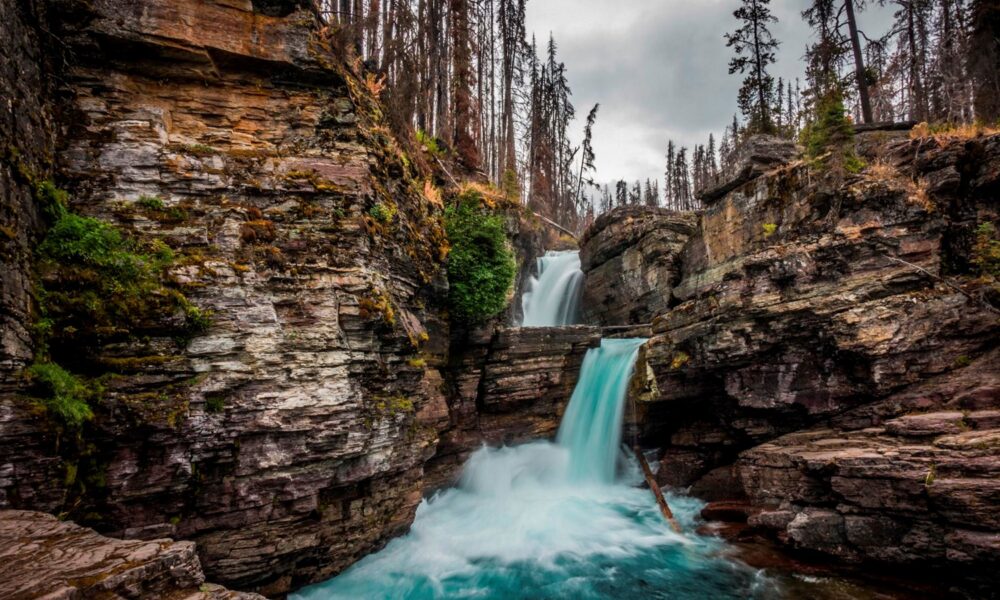 Real St. Mary Falls double waterfall dropping turquoise water between rocky cliffs, with forest and burned tree trunks in Glacier National Park