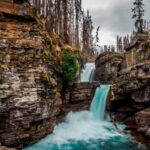 Real St. Mary Falls double waterfall dropping turquoise water between rocky cliffs, with forest and burned tree trunks in Glacier National Park